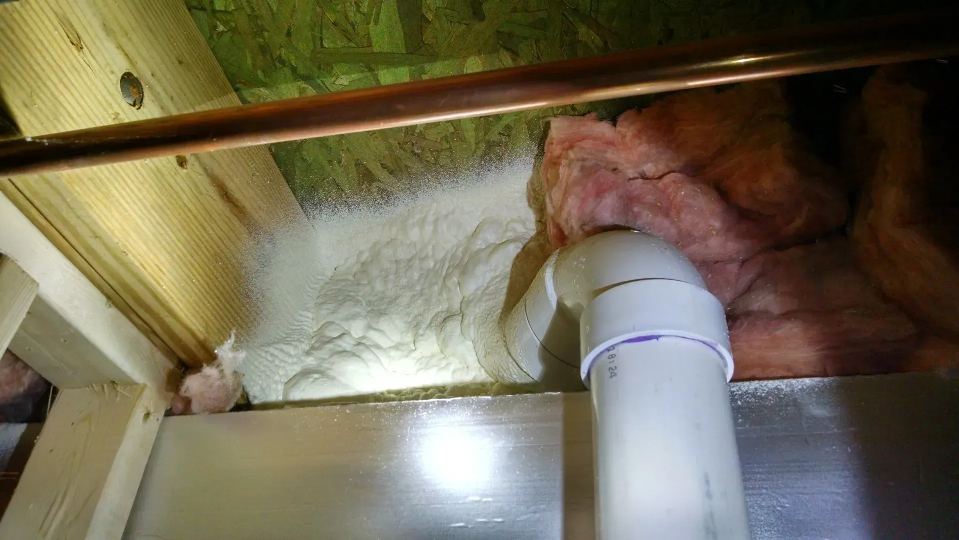 A PVC pipe passes through a wall cavity insulated with white spray foam and pink fiberglass, viewed from above.