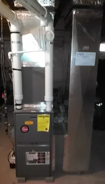 A grey Rheem furnace and an attached tall metal cabinet stand in a basement with white PVC venting pipes above.