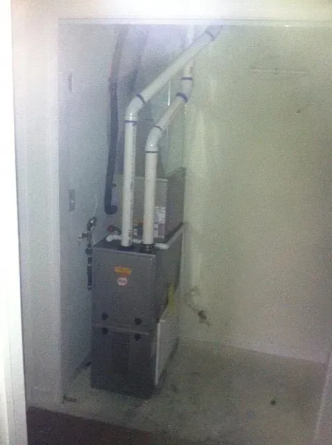 A grey HVAC furnace unit with white PVC intake and exhaust pipes installed in a small, white-walled utility closet.