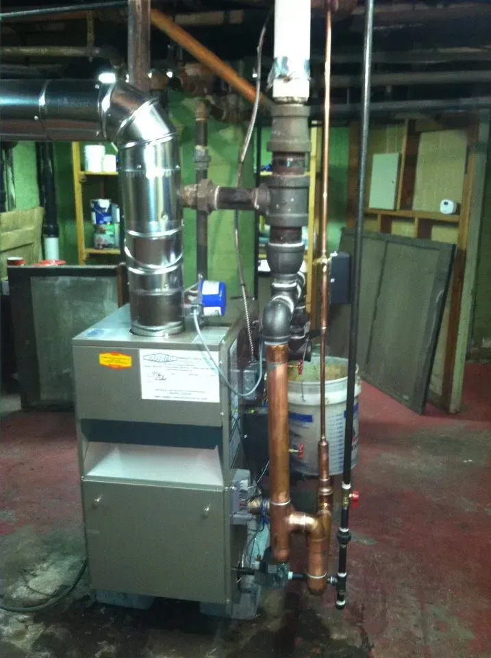 A metal furnace unit in a basement, connected to silver metal exhaust ducting and copper and black iron piping.