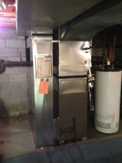A basement view of a metal furnace unit with a maintenance tag and an adjacent white water heater.