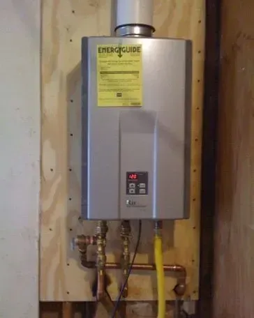 A silver tankless water heater mounted on a wooden board, featuring a digital display, yellow gas line, and plumbing.