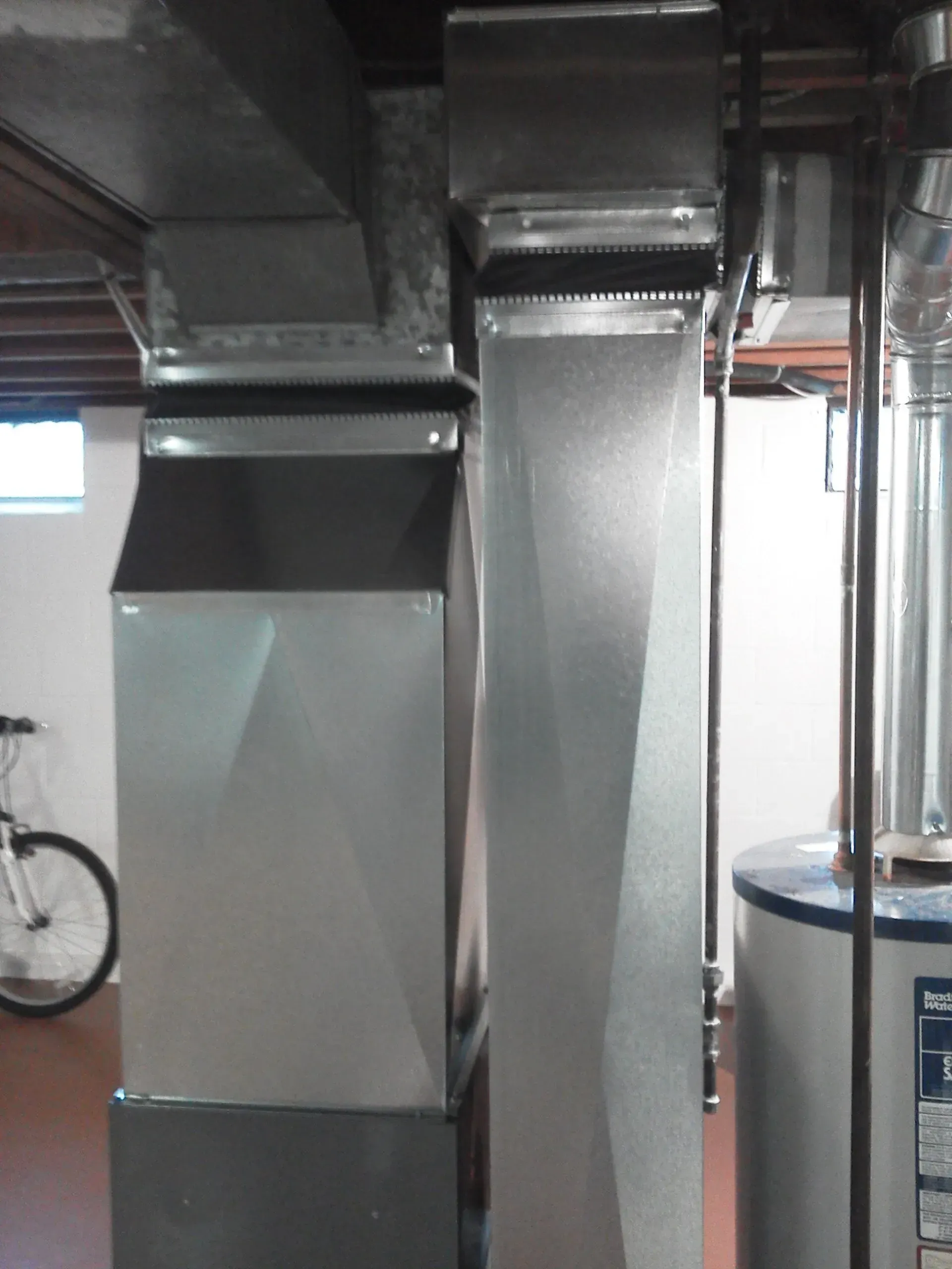 Two vertical metal ductwork sections attached to horizontal ceiling ducts in a basement setting.
