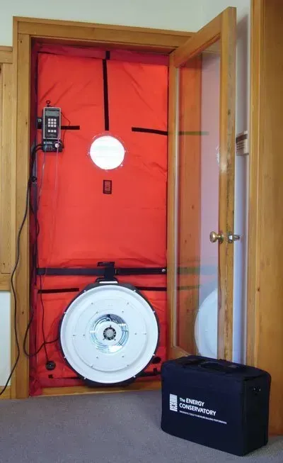 A blower door test setup with a red fabric panel, fan, and gauge mounted in a wooden doorway, with a carrying case nearby.
