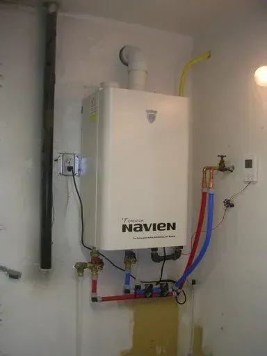 A white Navien tankless water heater mounted on a white wall with connected pipes and electrical wiring.