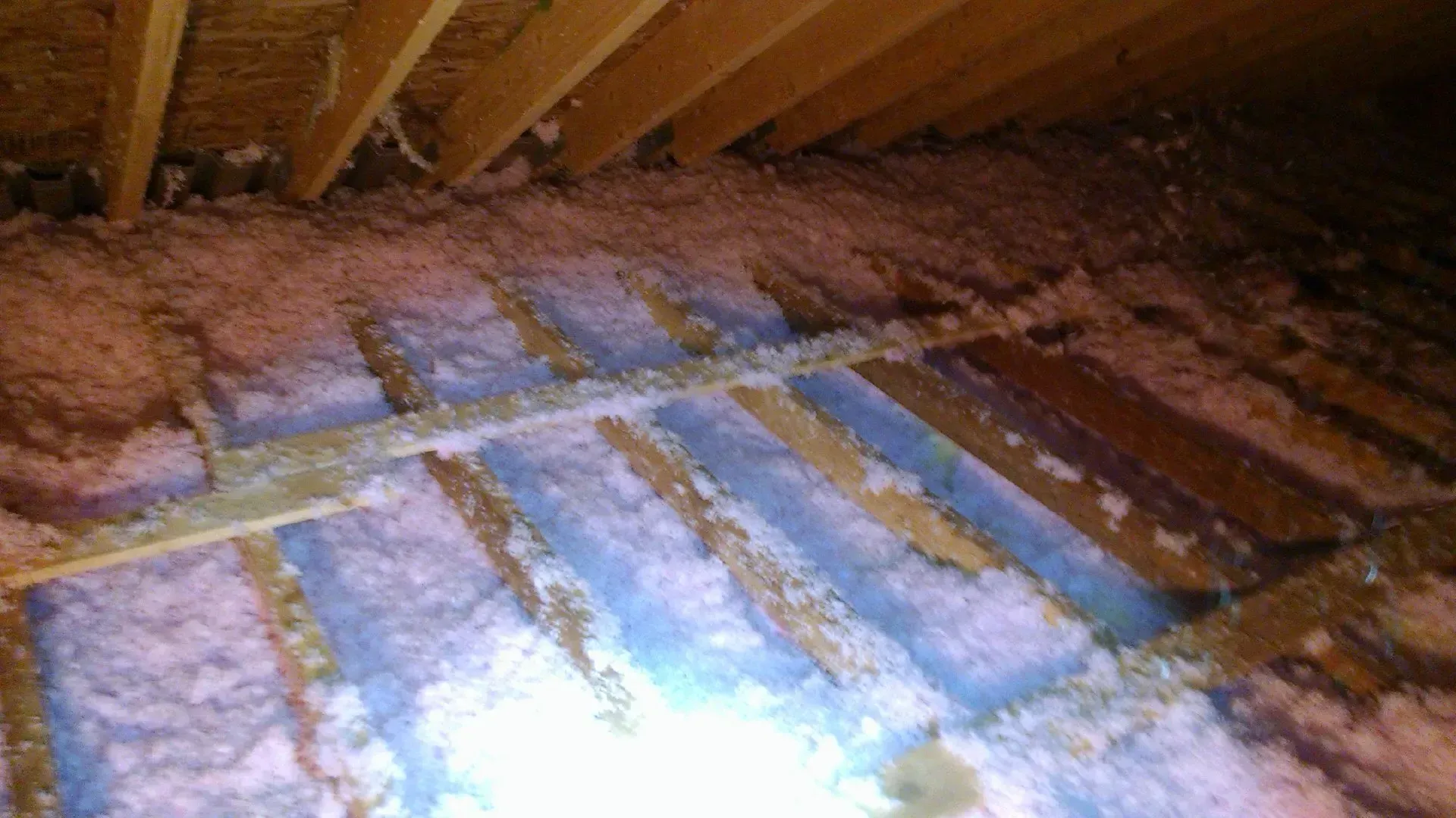 An attic floor featuring uneven pink and blue loose-fill insulation between wooden joists under a sloped roof.