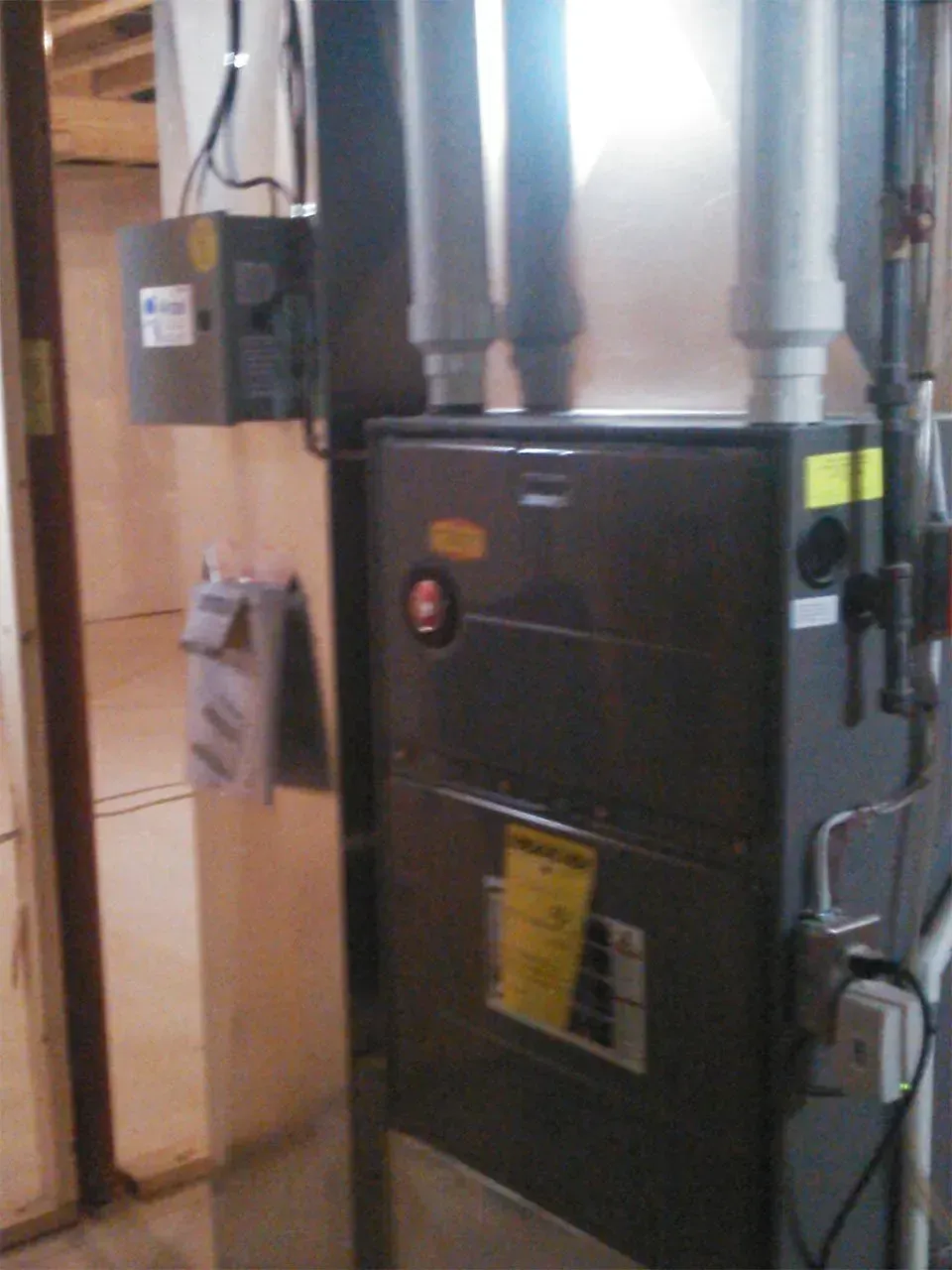 A dark gray furnace unit in a basement with two white vertical vent pipes on top and an electrical box mounted nearby.