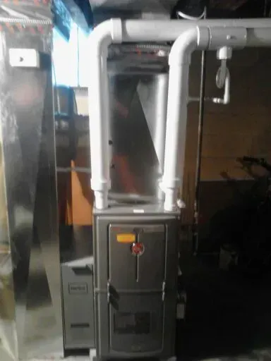 A gray gas furnace with two white PVC exhaust pipes attached to the top, situated in a basement setting.