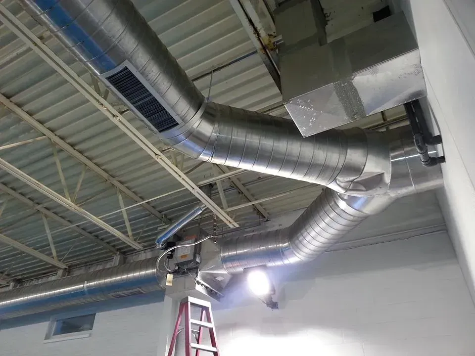 Silver industrial air ducts are mounted to the ceiling of a white room, with a red ladder visible beneath them.