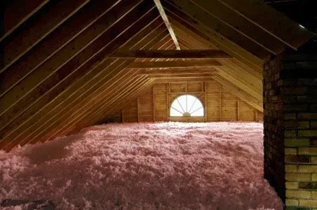 An unfinished attic with a semi-circular window, exposed wooden roof rafters, and a thick layer of pink insulation.
