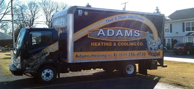 A box truck for Adams Heating & Cooling Co. parked on a street, featuring their company branding and phone number.