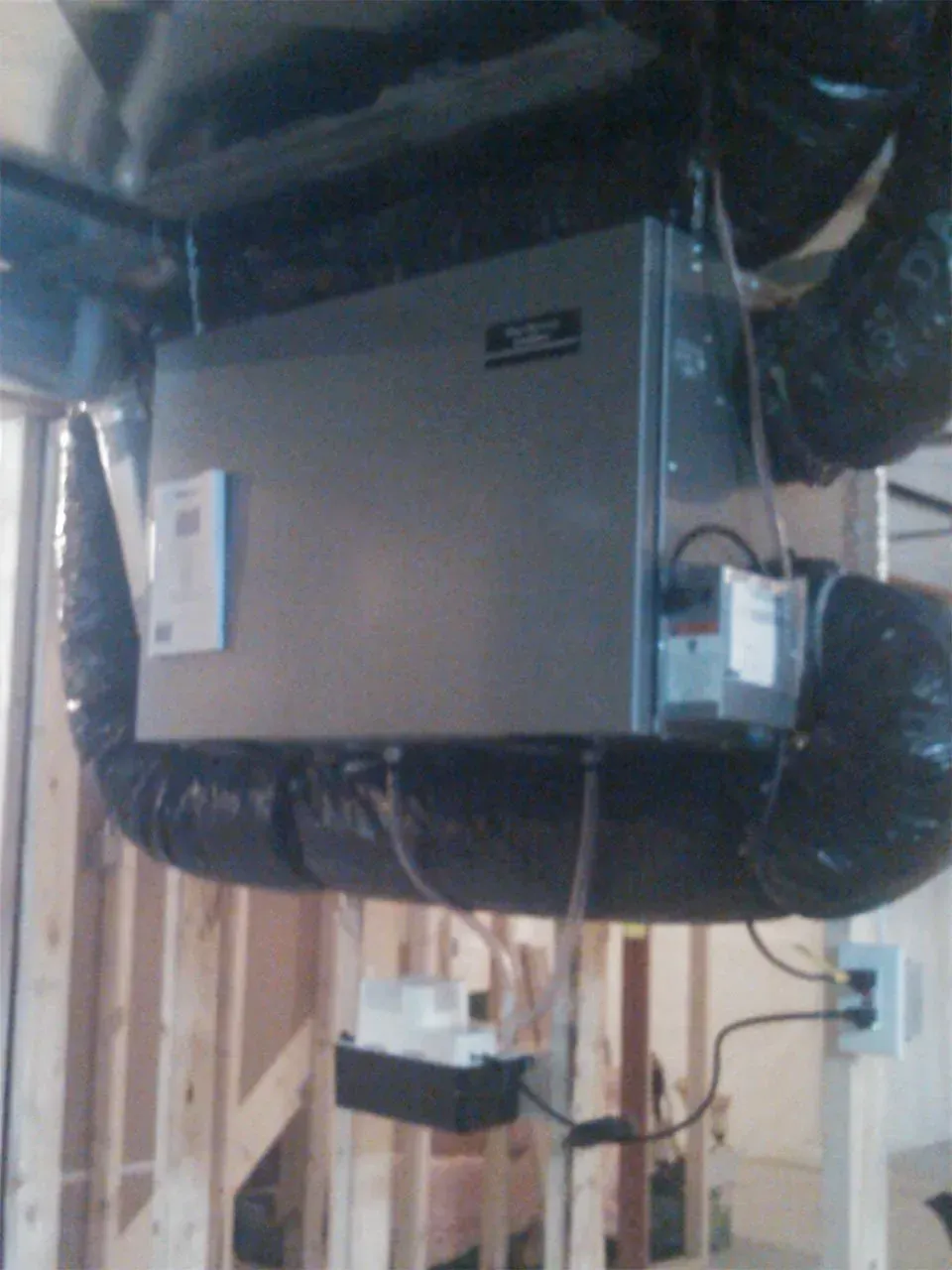 A grey HVAC unit hangs from ceiling joists in an unfinished basement, connected to black flexible ducts and a pump.