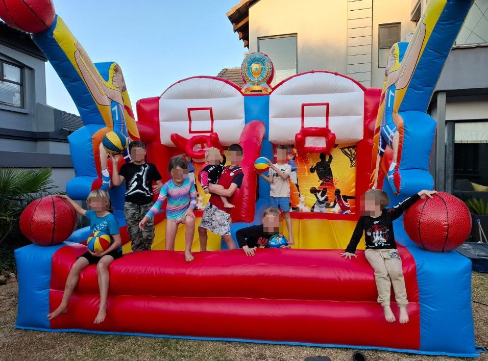 Children playing on a red, blue, and yellow inflatable basketball hoop bounce house outdoors.