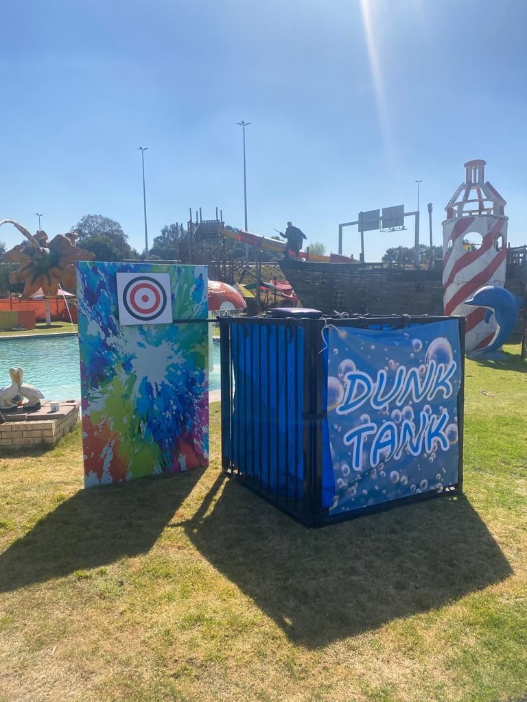 Dunk tank and target game on a grassy area, in front of a pool and amusement park. Bright, sunny day.