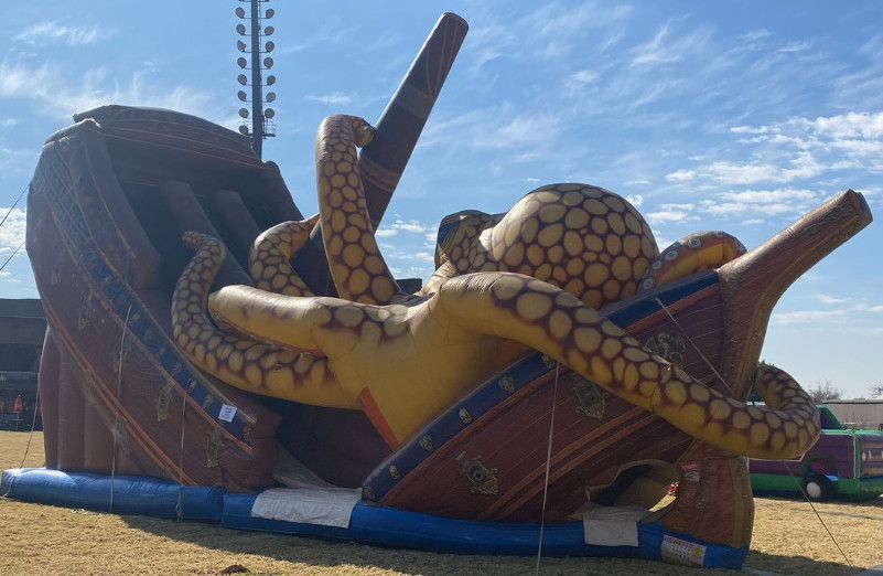 Giant inflatable octopus wrapped around a brown ship on a grassy field, under a blue sky.