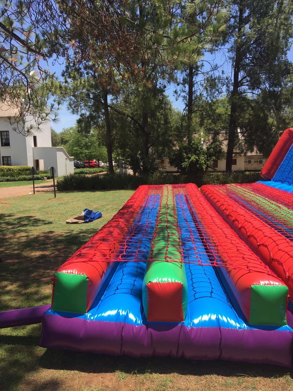Inflatable colorful obstacle course set up on a grassy lawn with trees and a white building in the background.