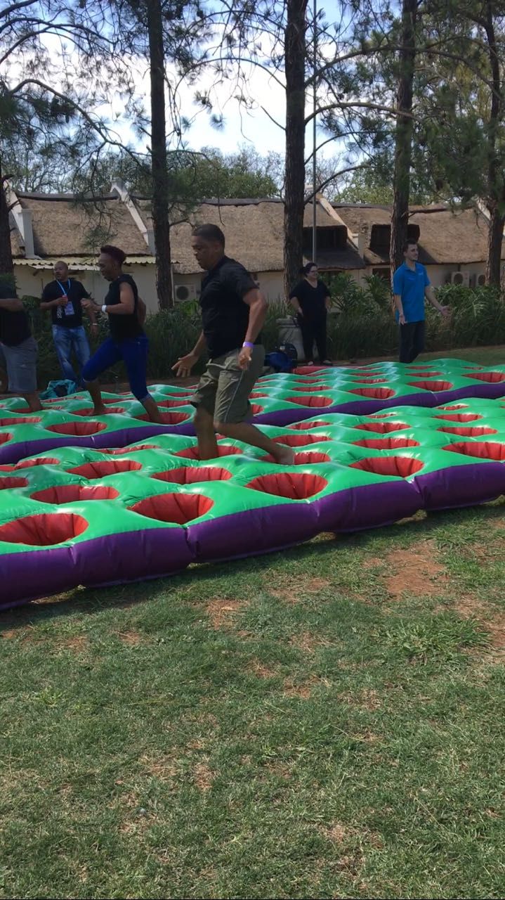 People jumping across an inflatable obstacle course with red and green sections in a grassy area.