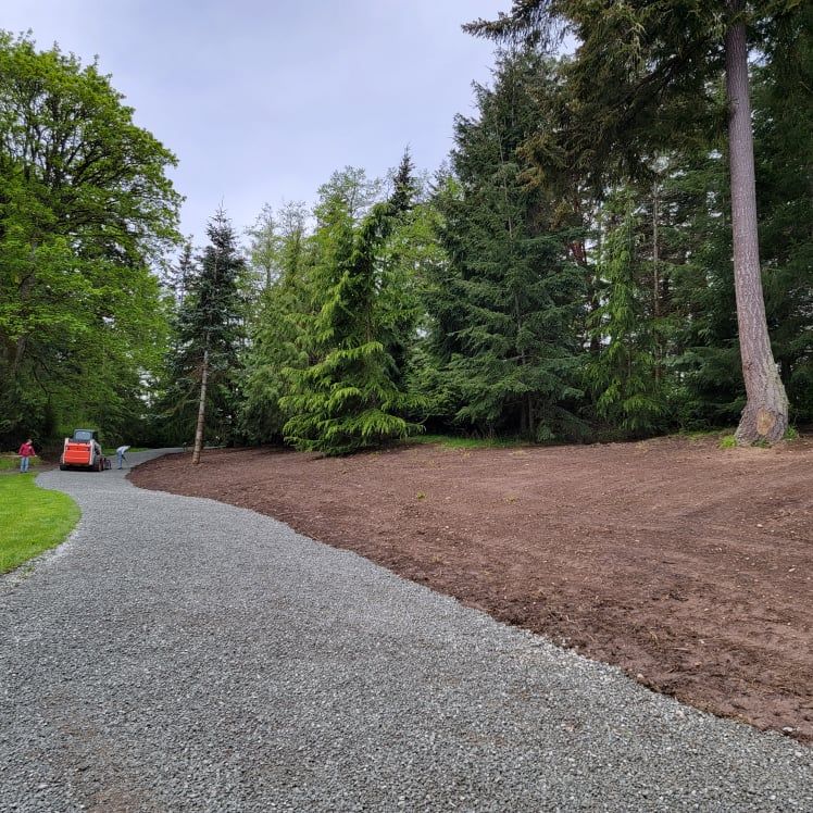 A smooth gravel driveway curves into a clearing next to a manicured tree line after excavation project has been completed. A small construction vehicle is present.