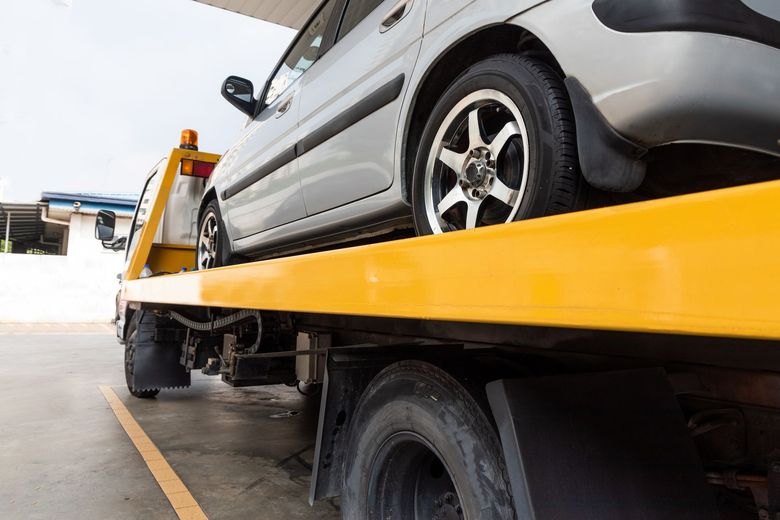 Silver car on a yellow tow truck bed, viewed from the side on a roadside.