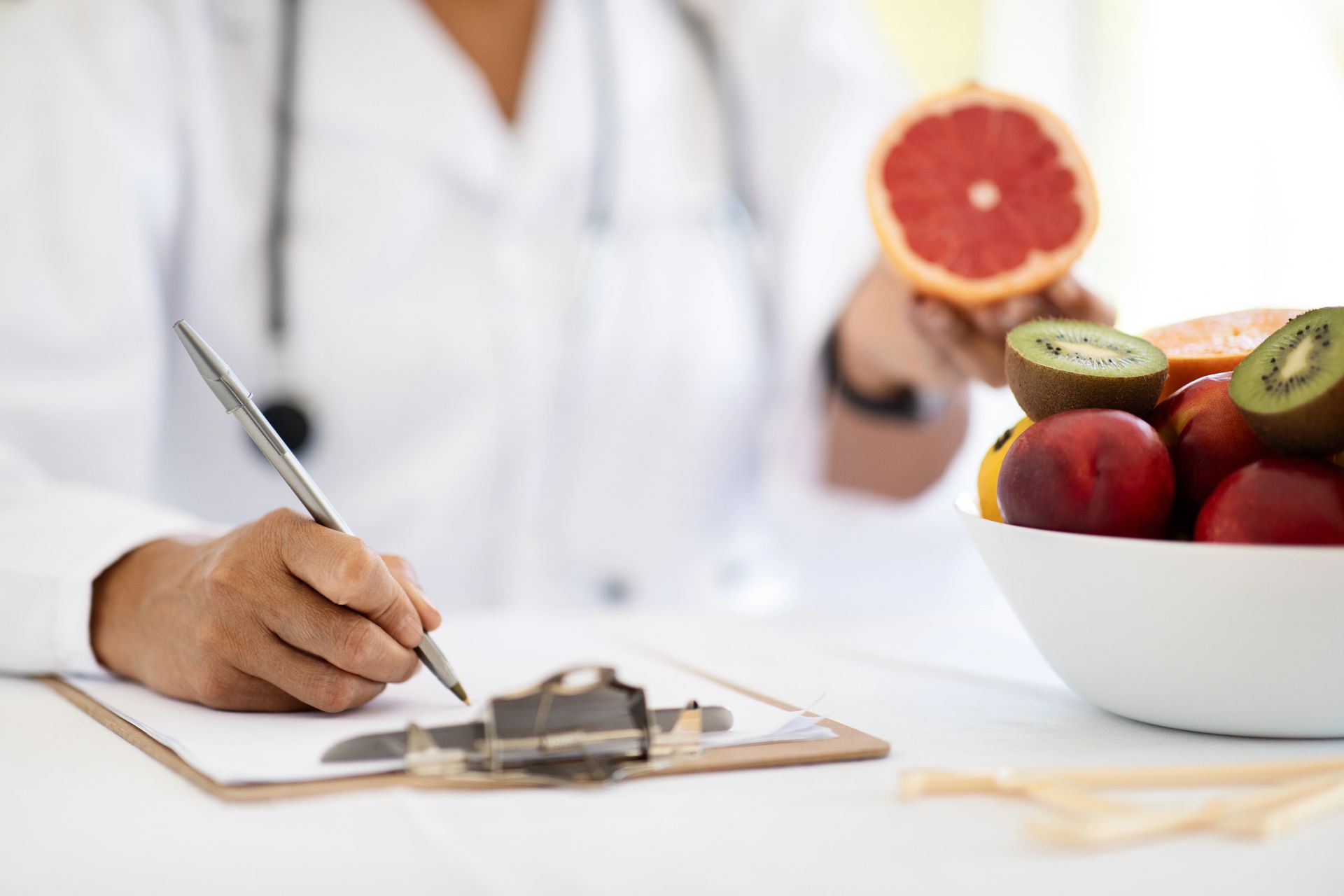 Medico che scrive su un blocco appunti, tenendo in mano un pompelmo. Una ciotola di frutta sul tavolo.