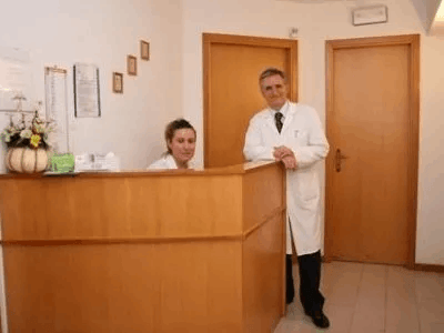 Receptionist e medico alla reception in legno di una clinica. La donna è seduta, l'uomo si sporge, entrambi sorridono.