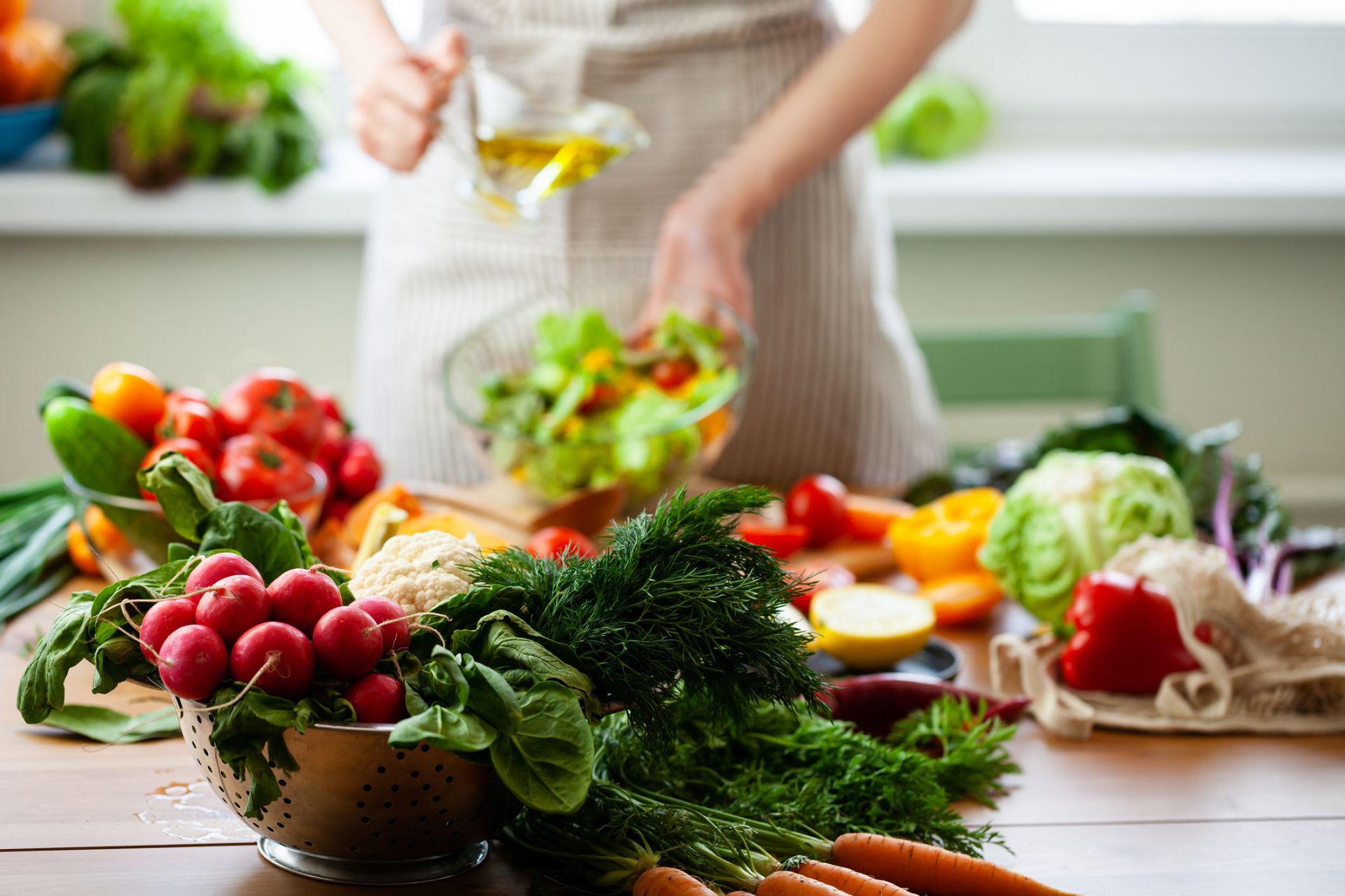Persona in cucina che versa olio sull'insalata; verdure fresche sul tavolo.