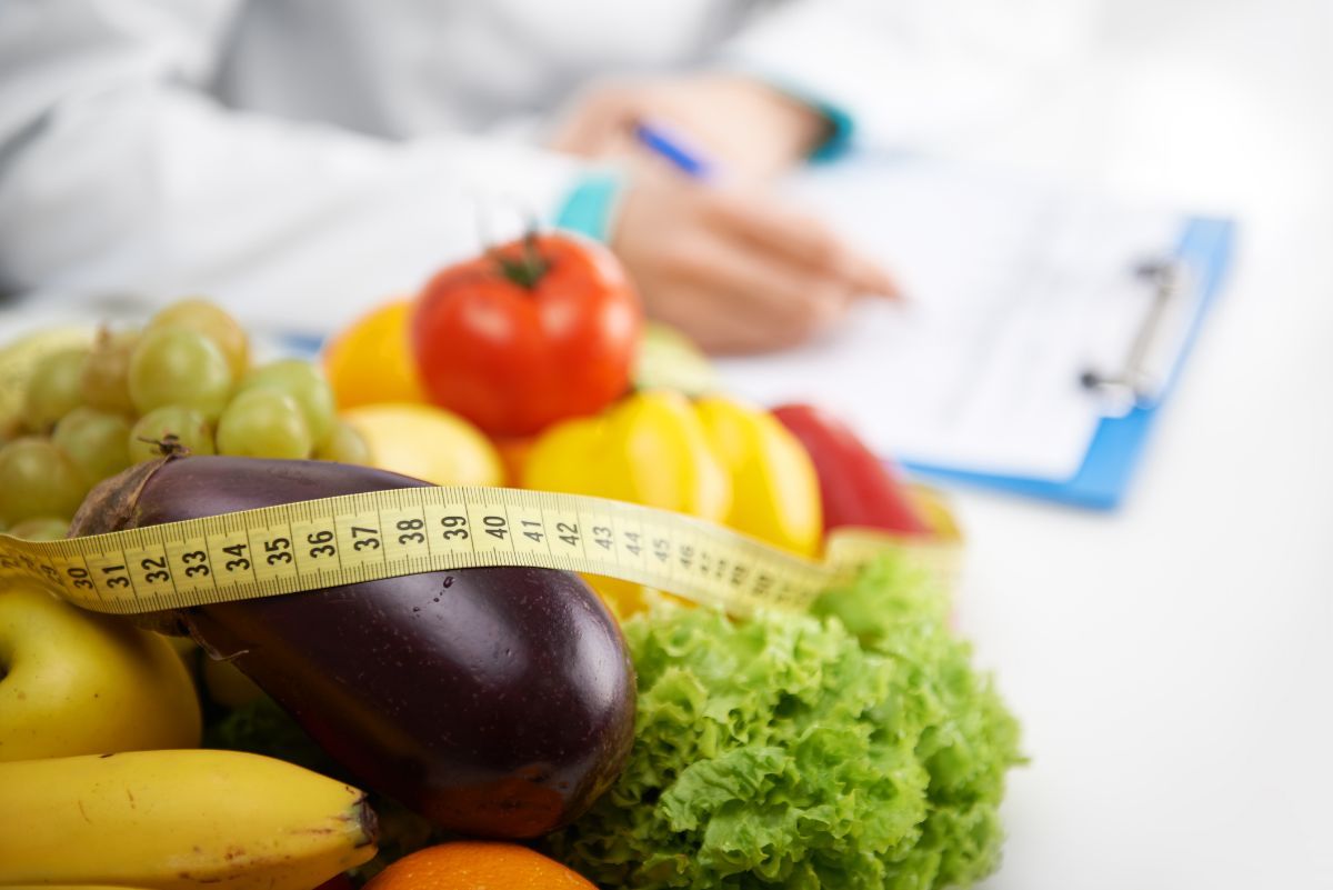 Frutta e verdura fresca con un metro a nastro; sullo sfondo un nutrizionista prende appunti su un blocco per appunti.