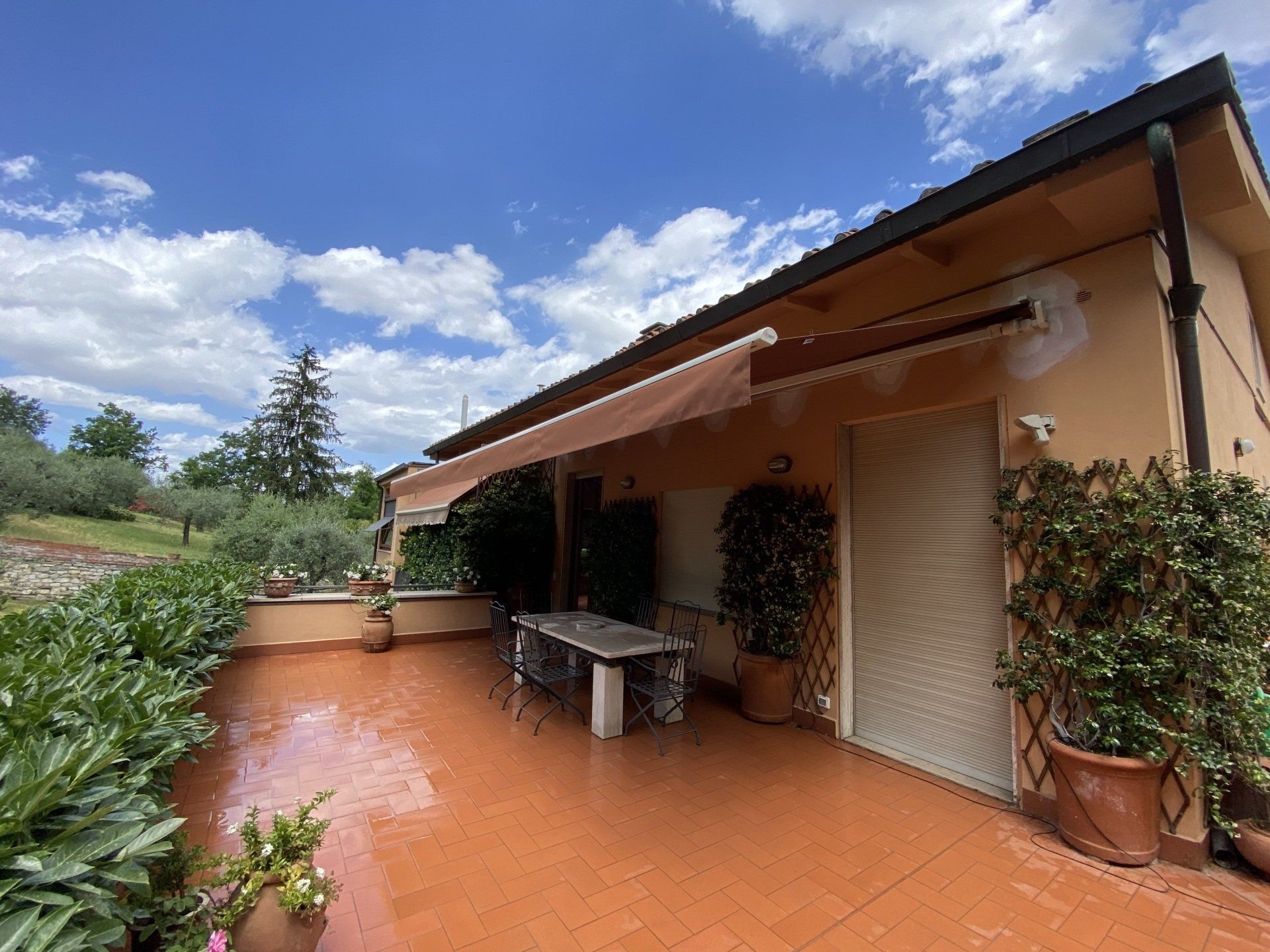 Terrazza con tenda da sole, tavolo, piante in vaso e cielo nuvoloso.