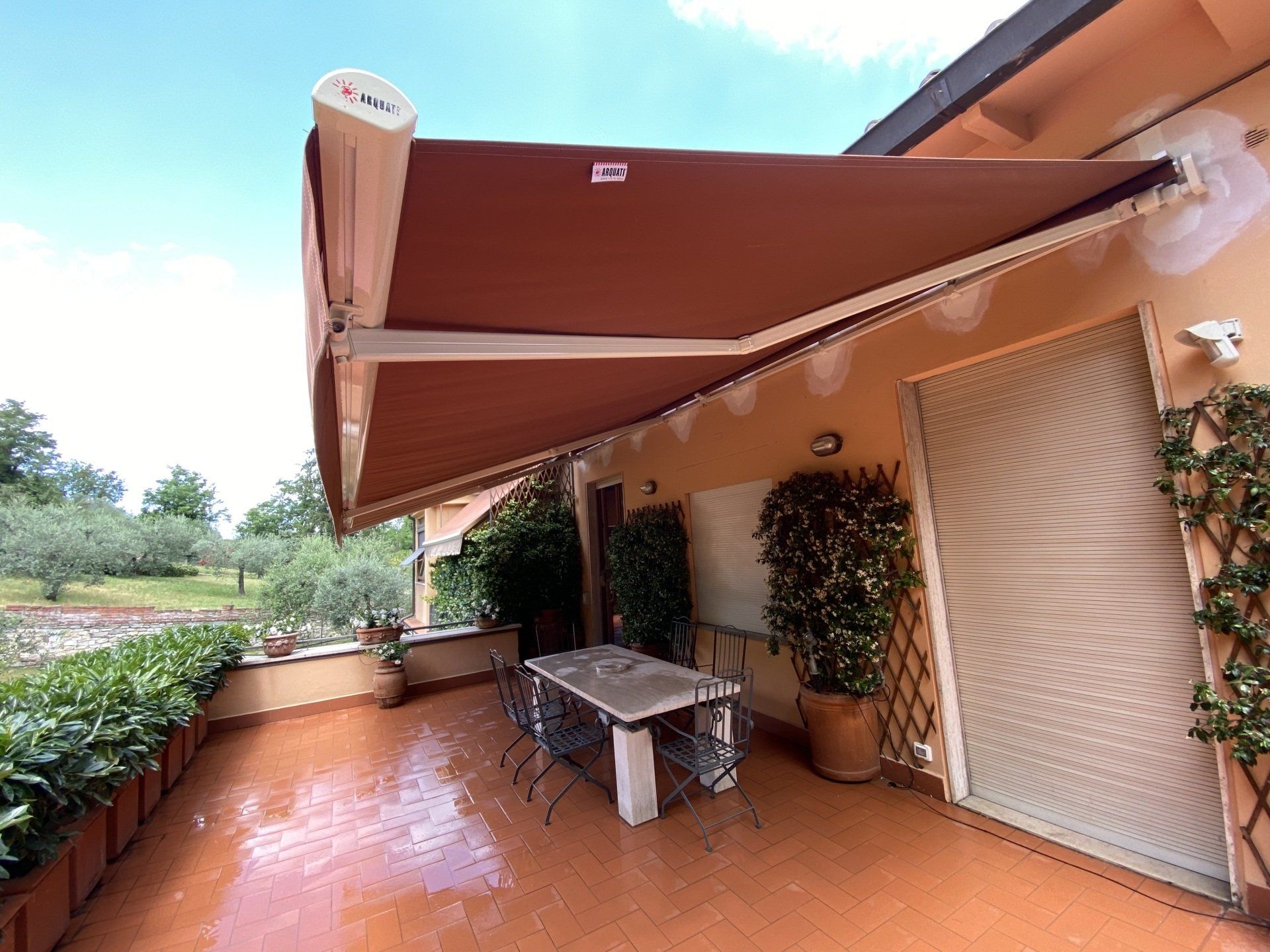 Tenda da sole marrone sopra una terrazza con tavolo e sedie, piante e vista sul verde.