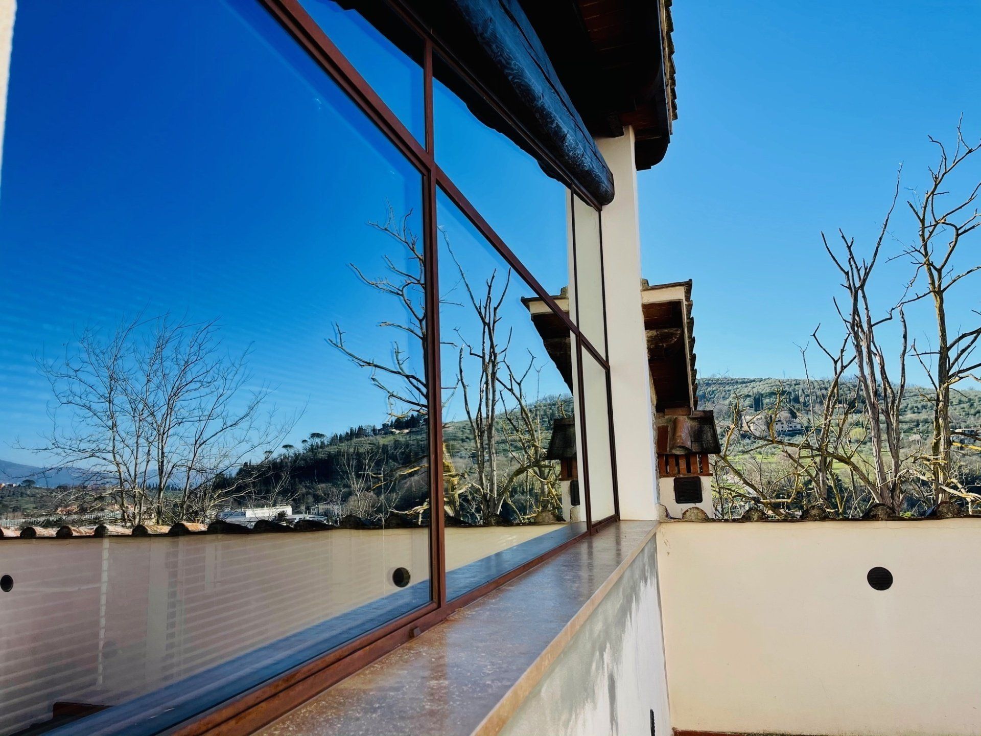 Una finestra che riflette un cielo azzurro brillante, con vista su un paesaggio collinare con alberi spogli e un muro bianco.