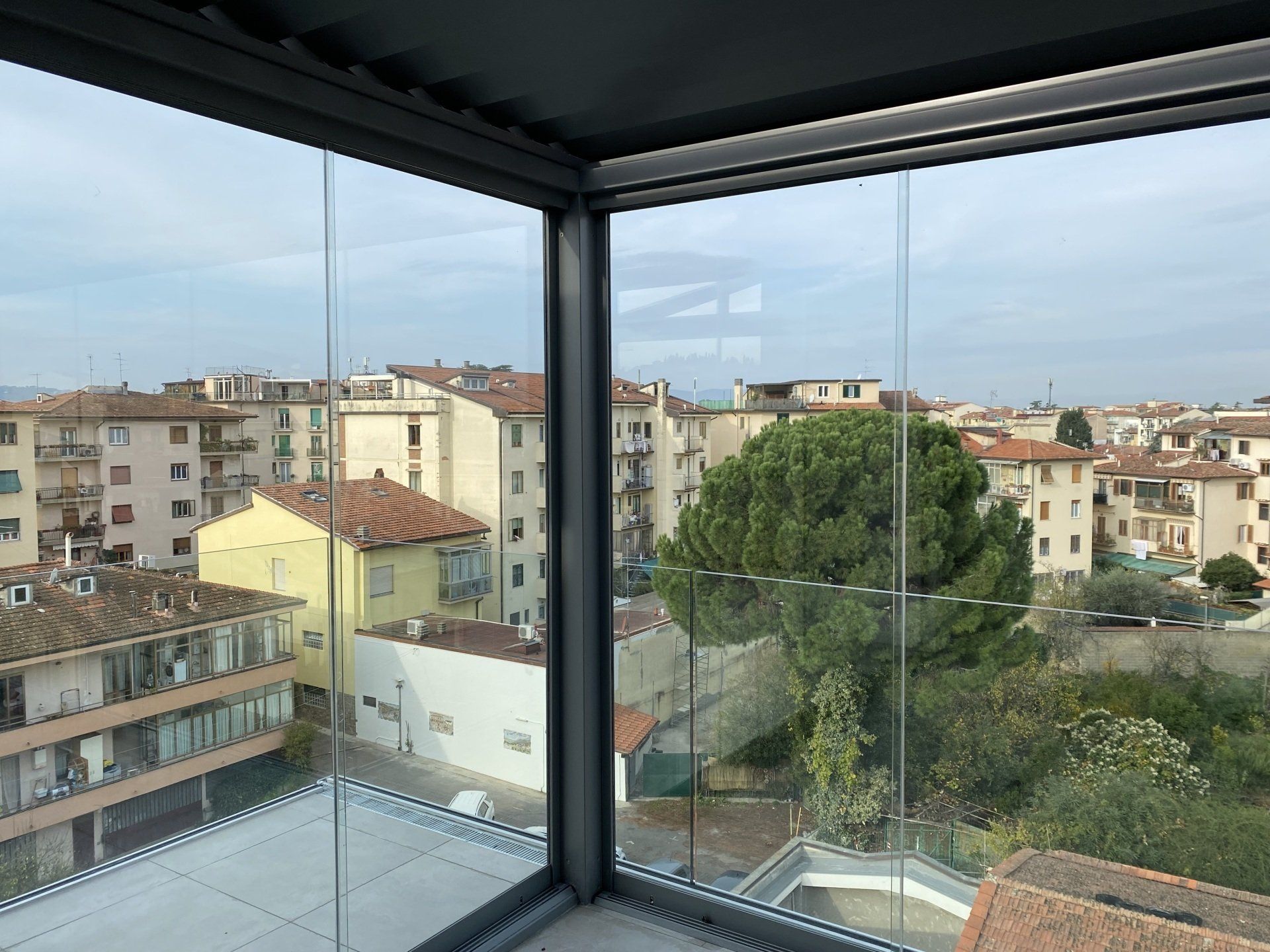 Balcone con pareti in vetro che si affaccia su un paesaggio urbano con edifici e un albero.