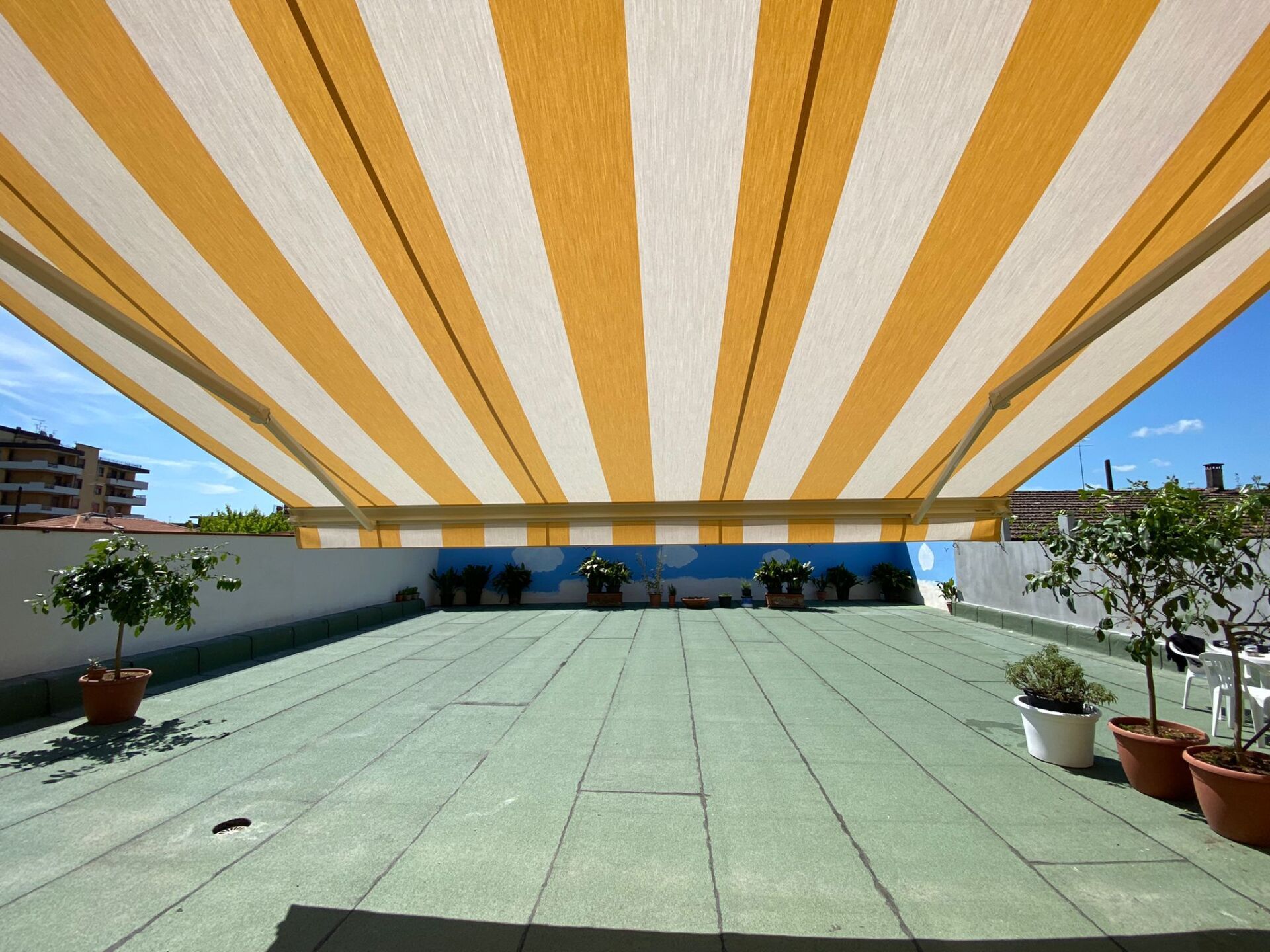 Una tenda a strisce gialle e bianche copre un patio sul tetto, con piante in vaso e cielo azzurro.
