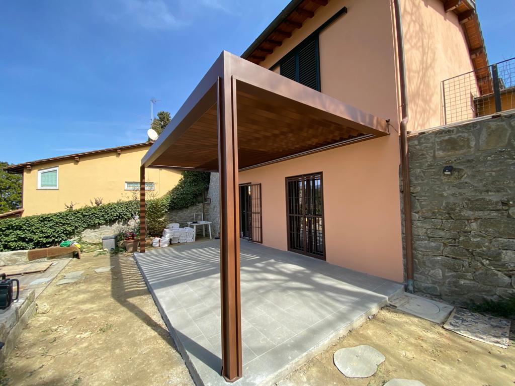 Pergola marrone che si estende da una casa color pesca su un patio in cemento. Giornata di sole.