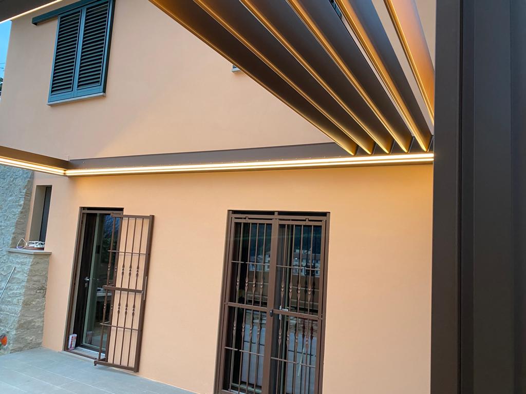 Un moderno pergolato marrone con luci integrate sopra un edificio con esterno arancione chiaro.
