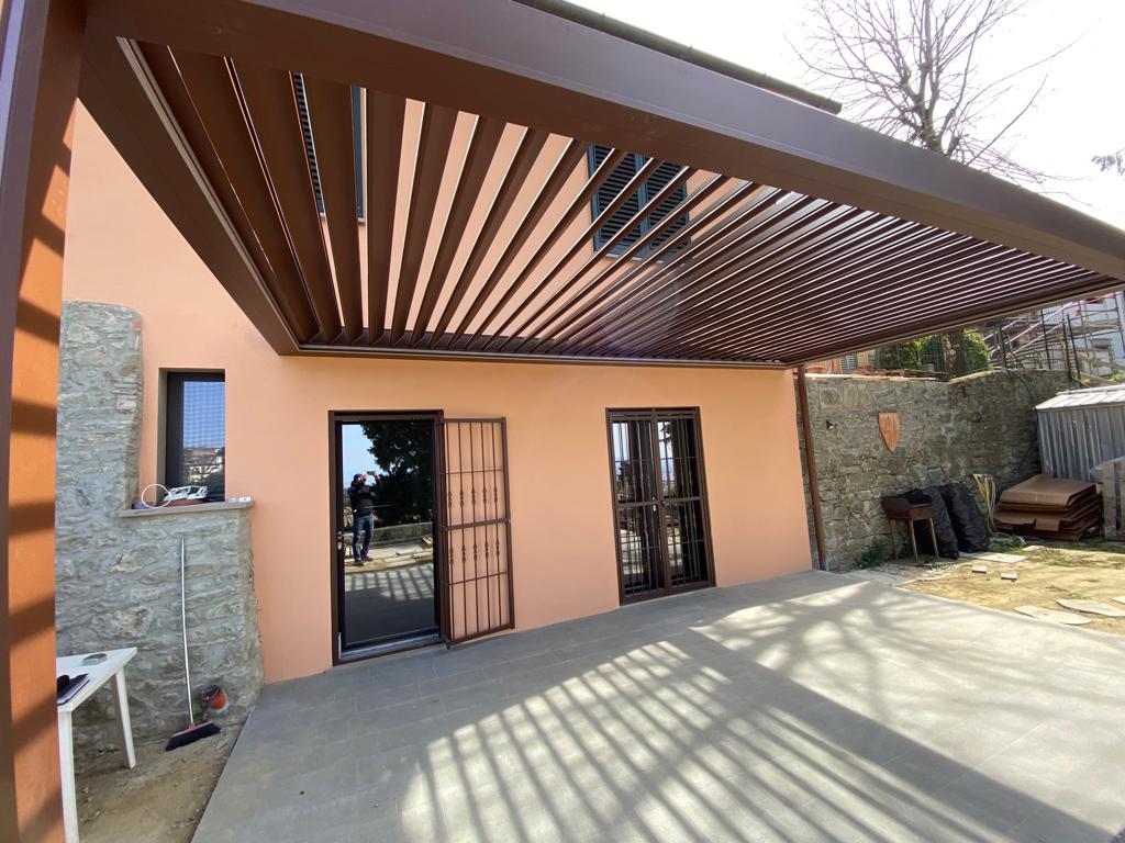 Pergola marrone sopra un edificio arancione chiaro con patio in cemento. La porta aperta mostra una persona.