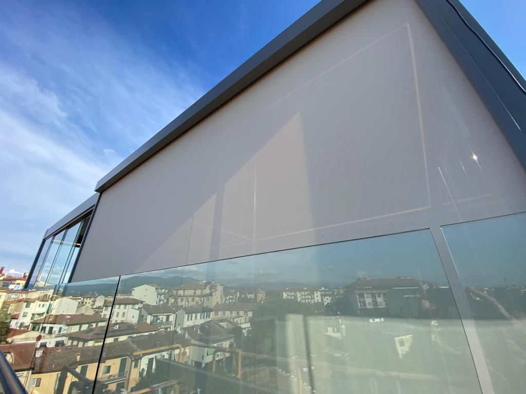 Tenda da sole beige su un edificio con ringhiera in vetro, con vista sulla città sotto un cielo azzurro.
