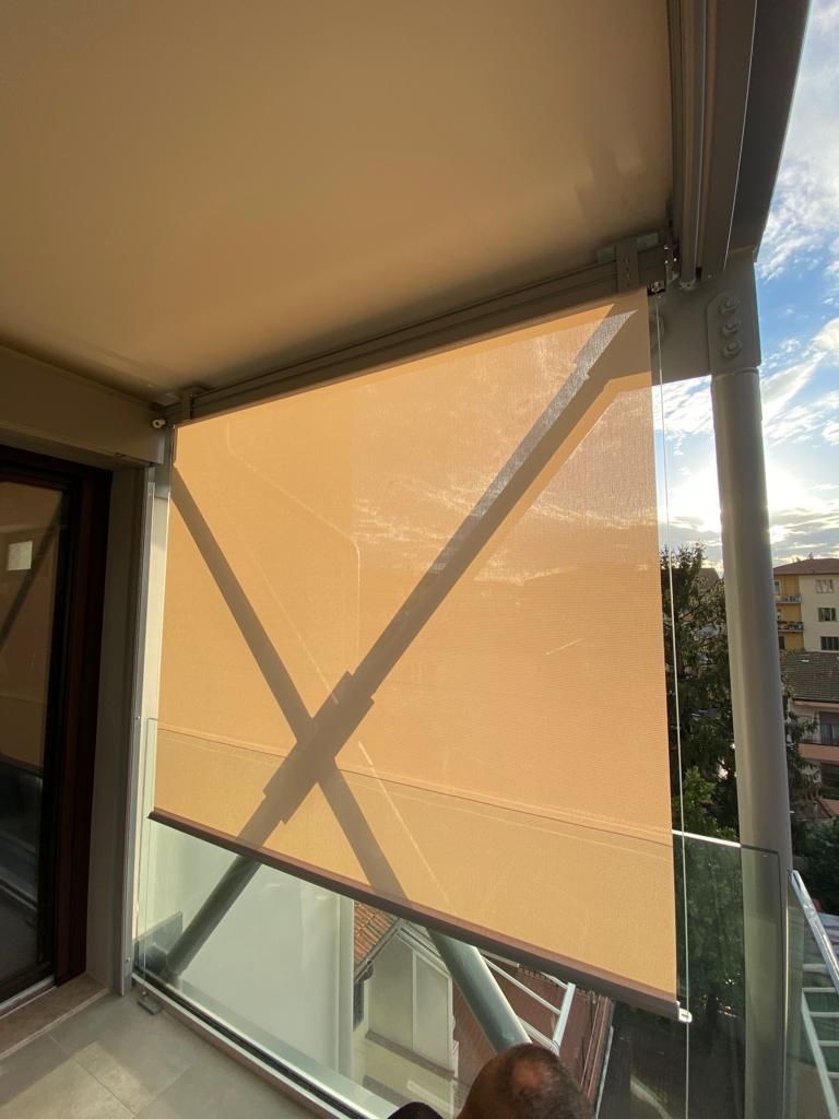 Tenda a rullo beige da esterno parzialmente abbassata su un balcone, contro una ringhiera in vetro e un cielo nuvoloso.
