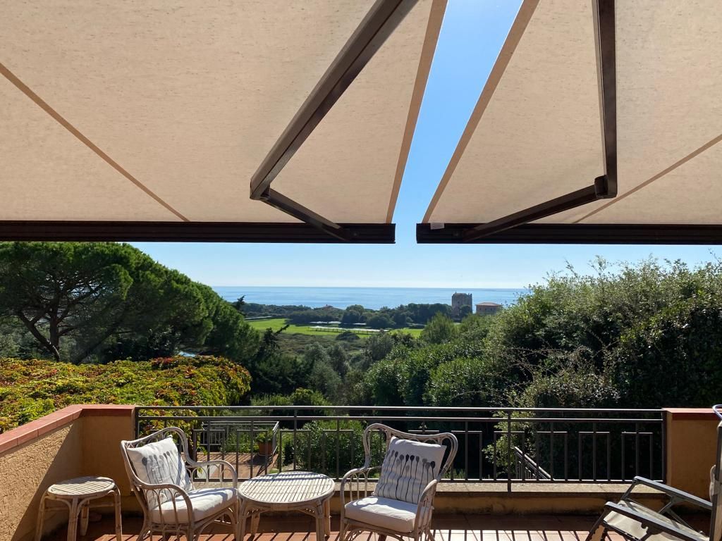 Patio con vista sull'oceano; tenda da sole con sedie e tavolo. Il cielo azzurro fa capolino; alberi e verde circondano.
