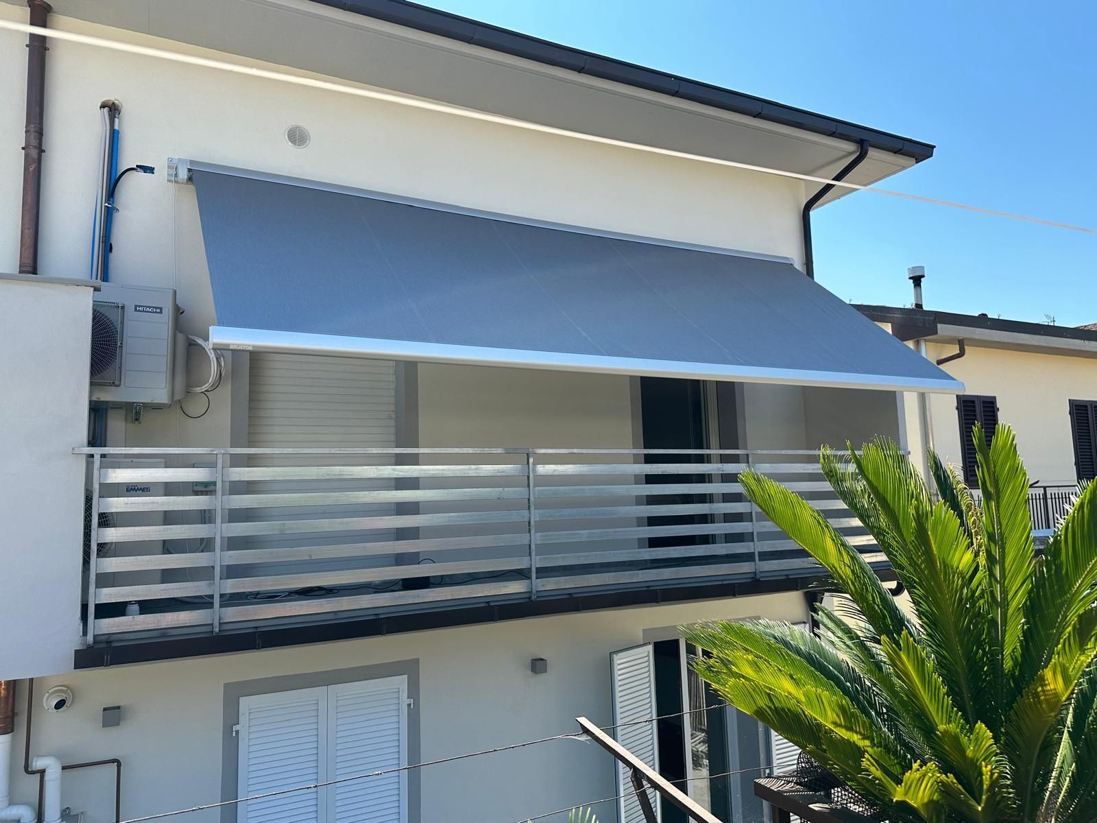 Tenda grigia su un balcone con ringhiera in vetro. Edificio bianco con aria condizionata. Giornata di sole.