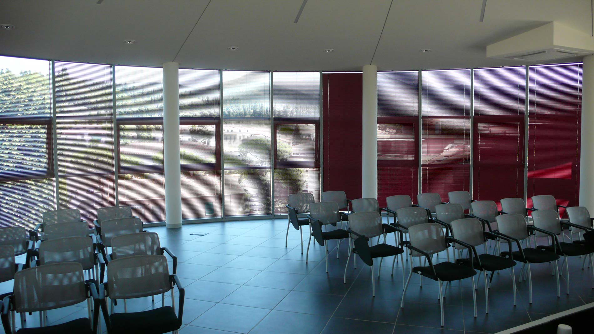 Sala conferenze con sedie di fronte alle finestre e tende a strisce. Dettagli color cremisi.
