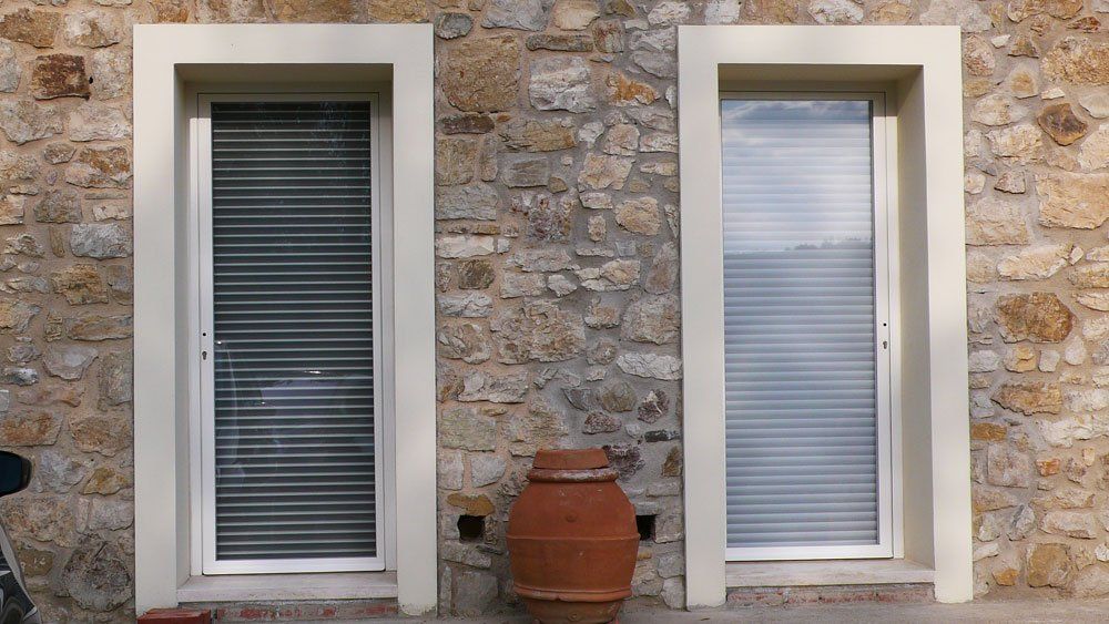 Due finestre con persiane chiuse nel muro di pietra, cornici beige. Sotto c'è un vaso di terracotta.