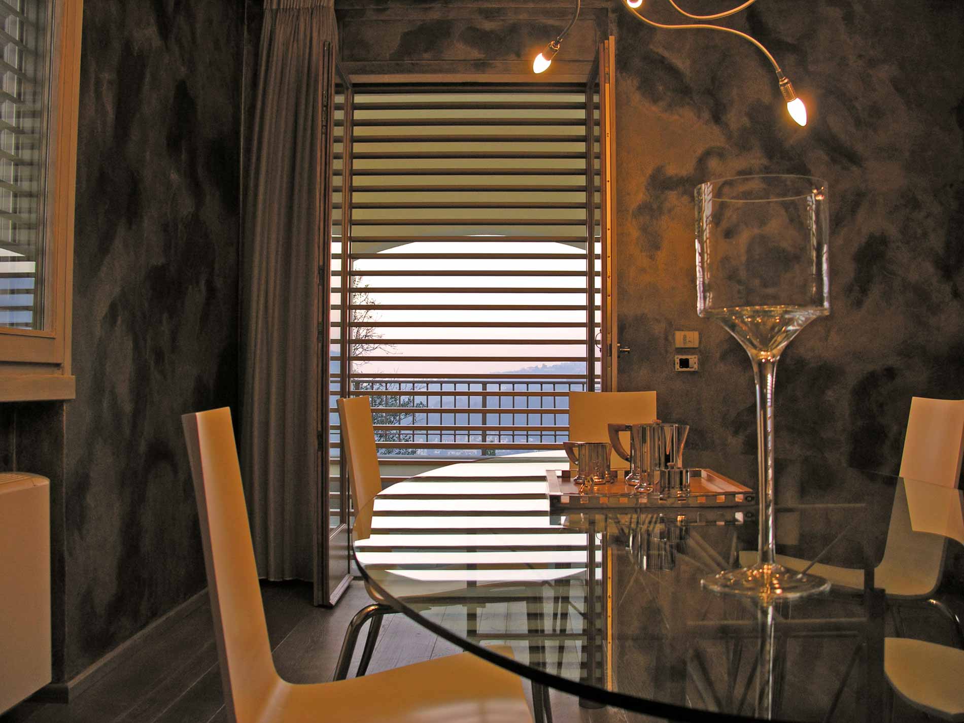 Sala da pranzo con tavolo in vetro, sedie e vista attraverso le persiane aperte.