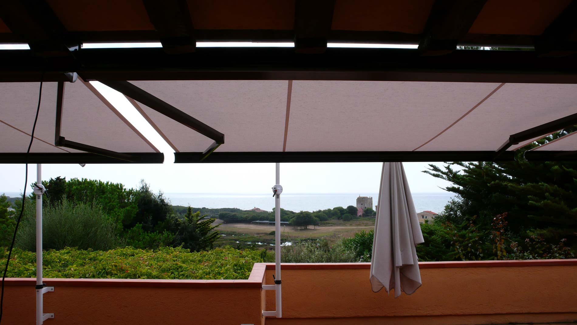 Patio con tenda da sole retrattile, vista sull'oceano, ombrellone bianco e vegetazione lussureggiante.