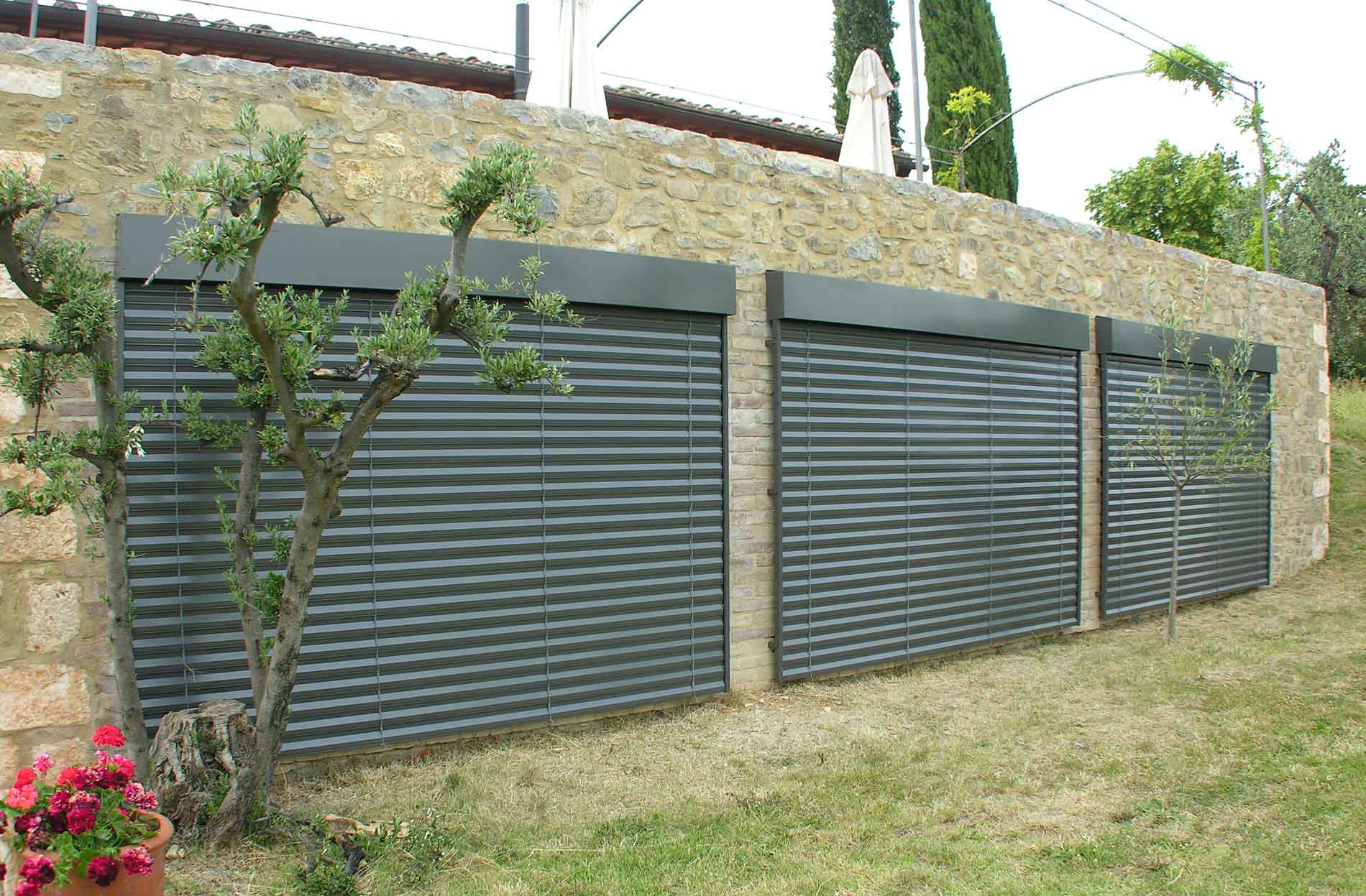 Tre tapparelle grigio scuro installate su un muro di pietra, in un contesto erboso.