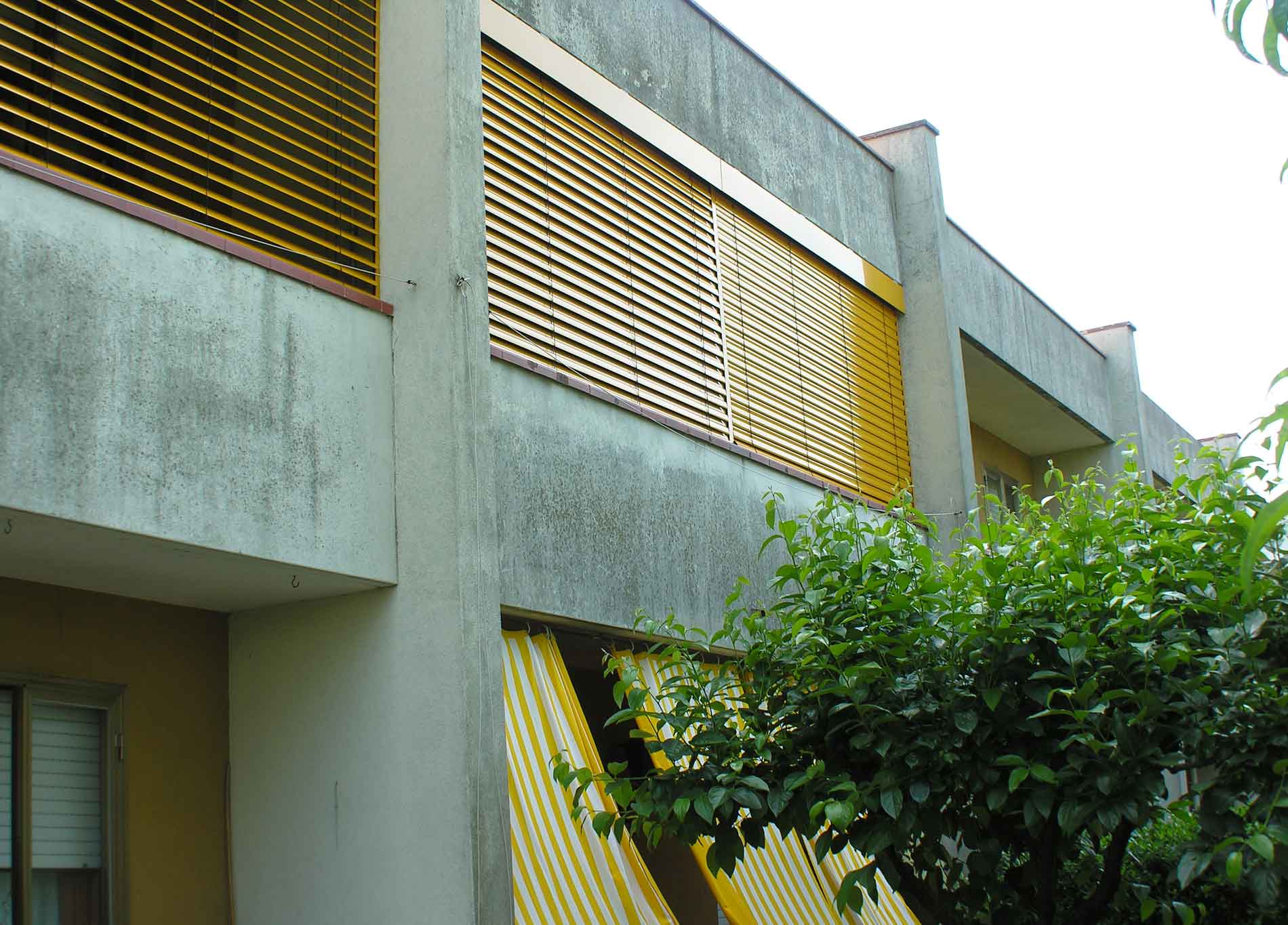 Esterno dell'edificio con tende a listelli gialli, cemento e un cespuglio verde.