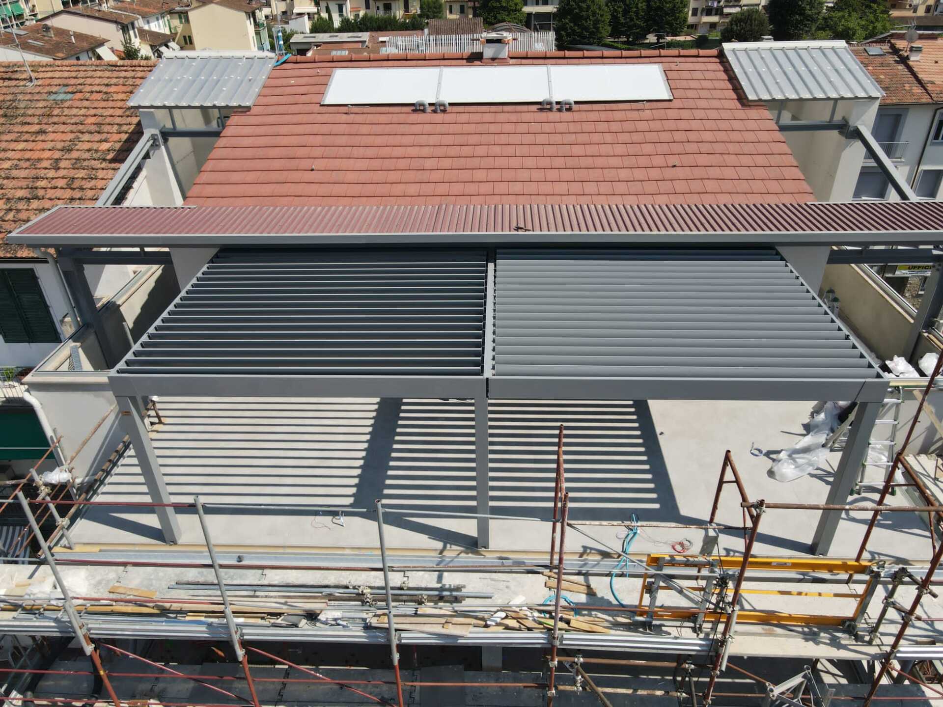 Pergola motorizzata grigia e nera su un tetto con un tetto di tegole rosse sullo sfondo.