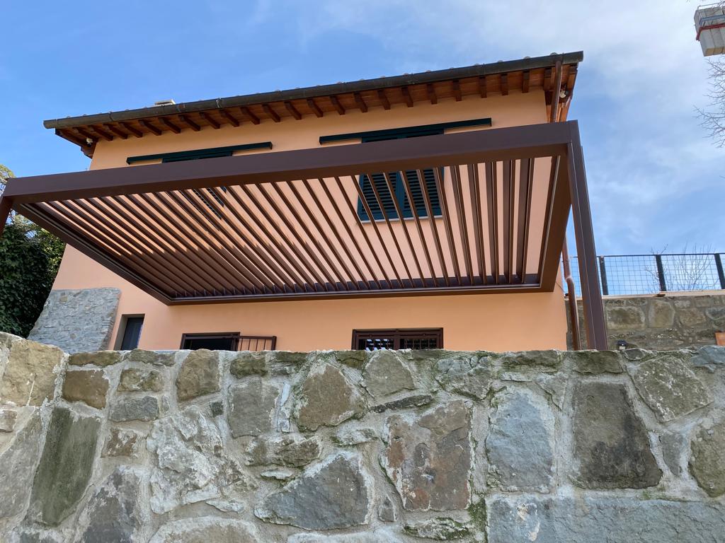 Pergola marrone sopra una casa color pesca con un muro di pietra in primo piano e un cielo azzurro sopra.