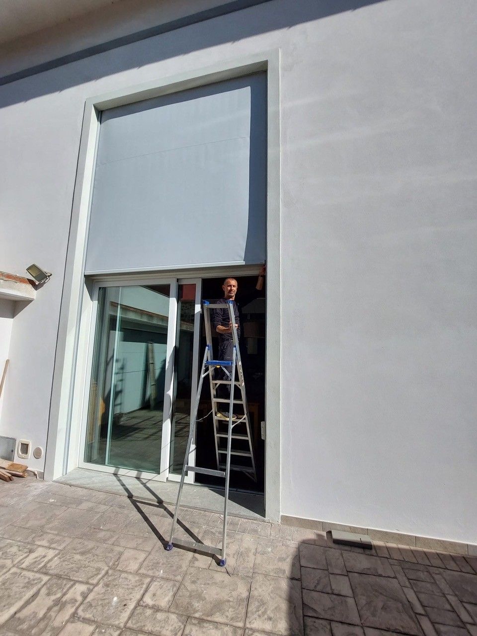 Uomo su una scala che installa una grande tenda esterna grigia sopra le porte scorrevoli in vetro di un edificio bianco.