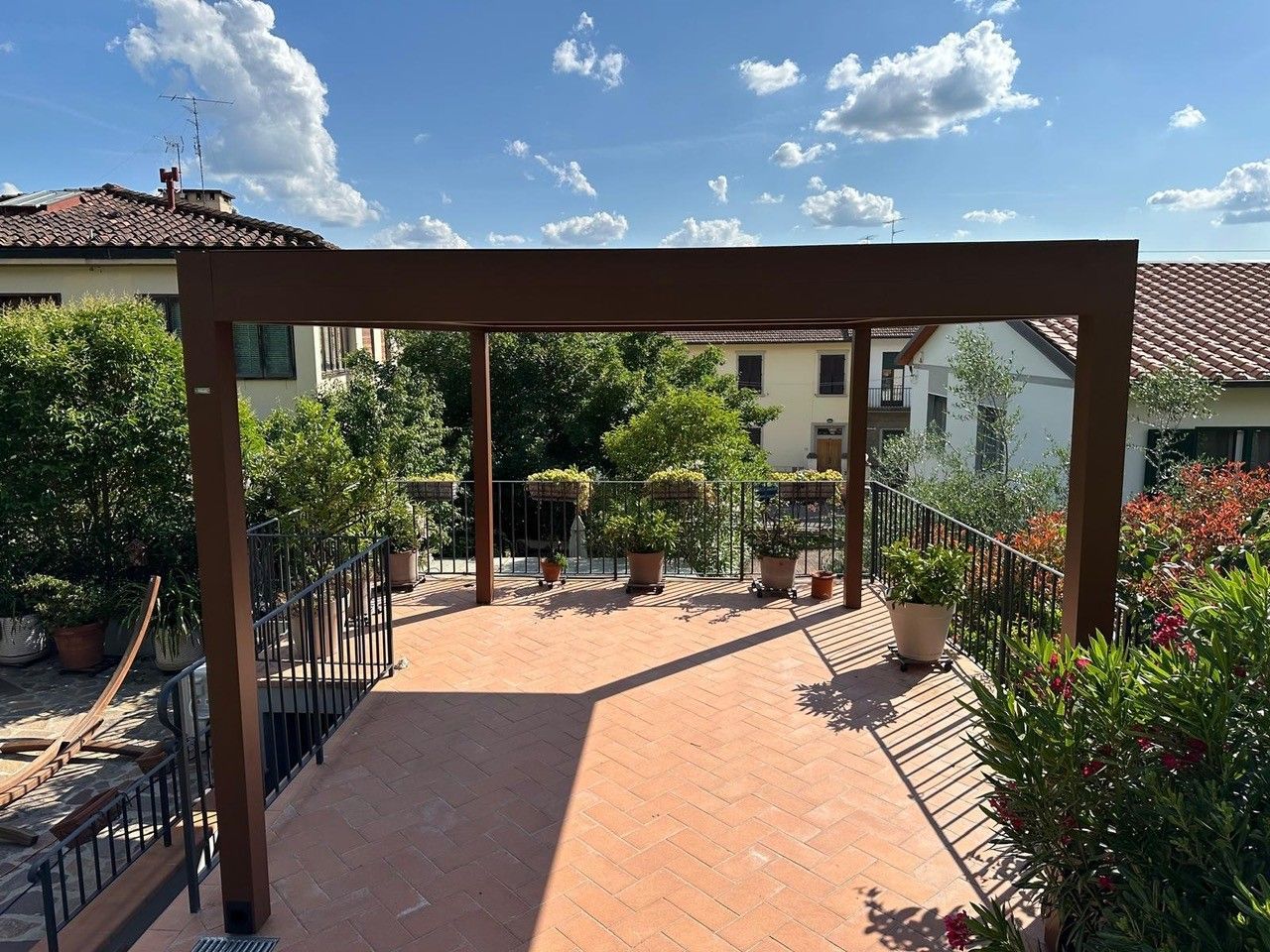 Pergola su un patio in terracotta, circondata da piante in vaso e da una ringhiera, contro un cielo soleggiato.