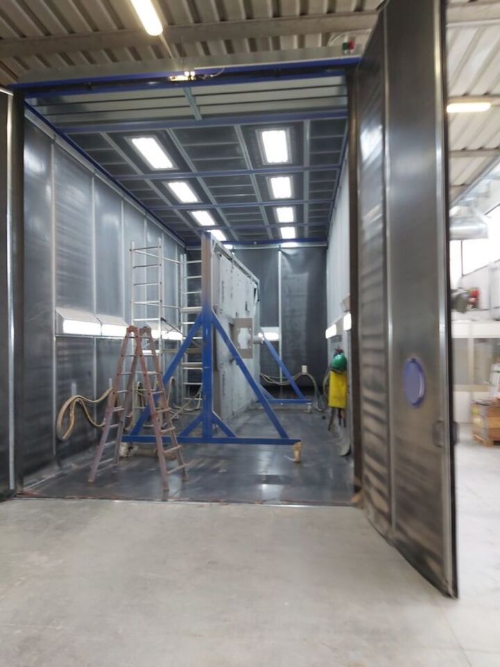 Cabina di verniciatura industriale aperta con attrezzature blu, avvolgitubo e luci intense dall'alto.